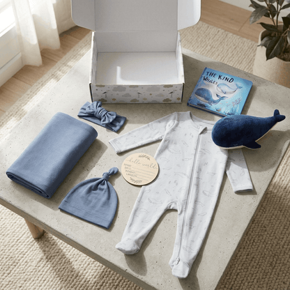 A baby gift set arranged on a light stone table, including a white footed sleeper with a subtle print, a folded blue blanket, a blue knit hat, a blue bow headband, a blue whale plush toy, an open gift box, and a children's book.