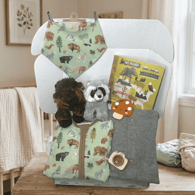 A baby gift set arranged on a white chair in a softly lit nursery. The set includes a light green bib and matching footed pajama set with woodland animal illustrations, a gray long-sleeve shirt, a yellow 'An Aardvark Adventure' book, two plush toys, and a small mushroom-shaped accessory.