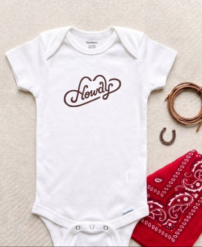 White baby onesie with 'Howdy' print on a textured surface with bandana and horseshoe.